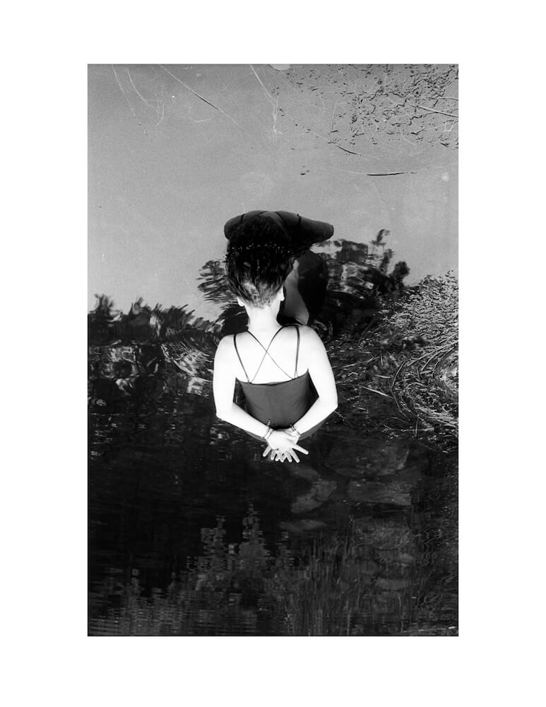 an upside down scan of a 35mm black and white negative of a woman in a dress dipping her hair into a lake